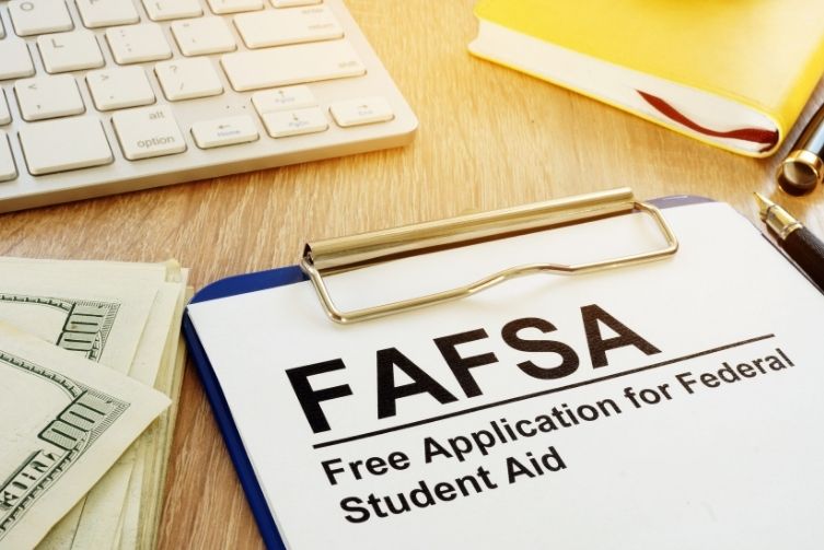 A clipboard holds a paper with the words FAFSA Free Application for Federal Student Aid with a stack of hundreds on the left side of the frame and a keyboard on the top left.