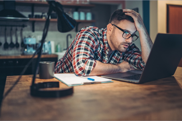 A man leans toward his desk with his hand on his head, as he stares at his laptop.