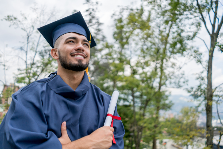 A young man wearing a graduation gown and hat smiles with his arms crossed as he holds his diploma.