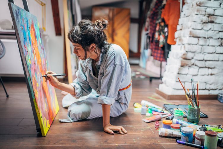 A woman paints on a canvas with art supplies around her.