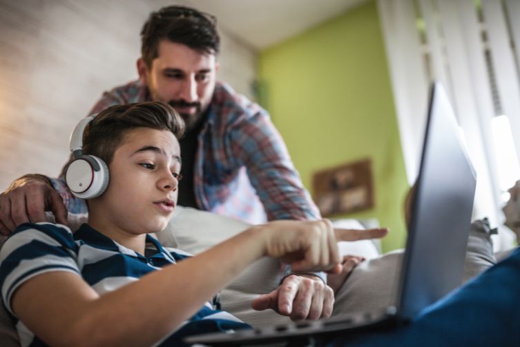 A young boy works on a laptop as his father looks over his shoulder.