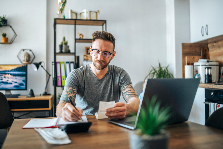 A man with glasses sits at a table with his laptop to handle his finances