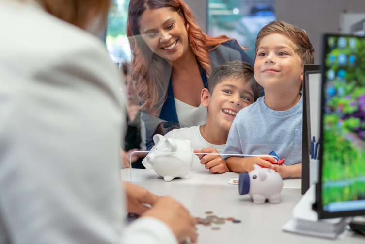A woman and her two young boys smile as the teller's arm can be seen sorting through the coins in their piggy banks in the foreground