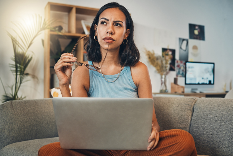 A young woman sits on a sofa with her laptop. She holds the tip of a pair of glasses to her chin as she looks pensive.