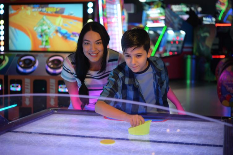 A woman watches her young son play air hockey at an arcade.