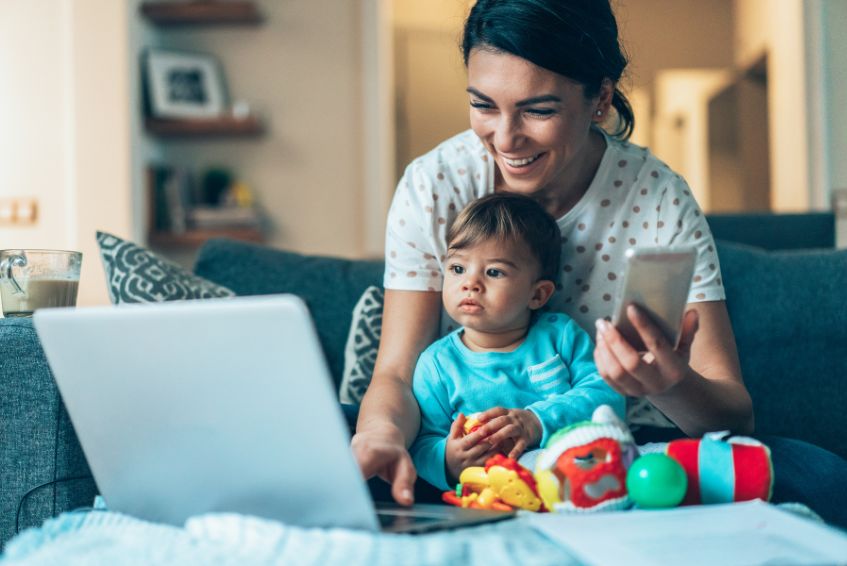 Woman, with baby in lap, holds phone in her hand and smiles at her laptop