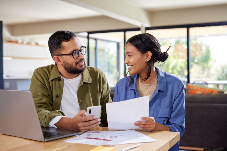 A man and woman sit at a table with a laptop and statements to discuss finances.