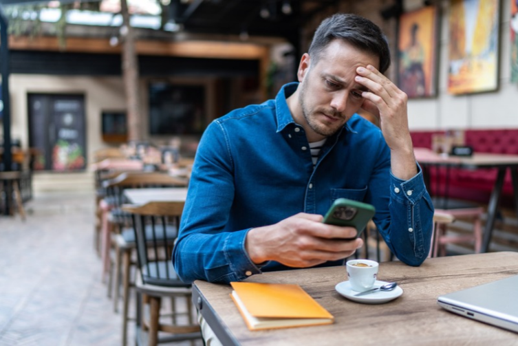 A man in a blue shirt sits worried at a coffee shop as he looks at his phone.