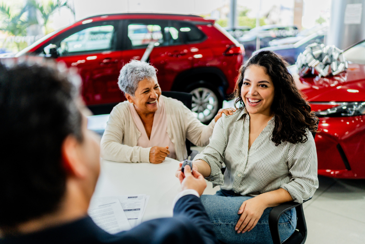 A young woman smiles as she's handed the keys to a new car. Her grandmother smiles with her hand on her granddaughter's shoulder.