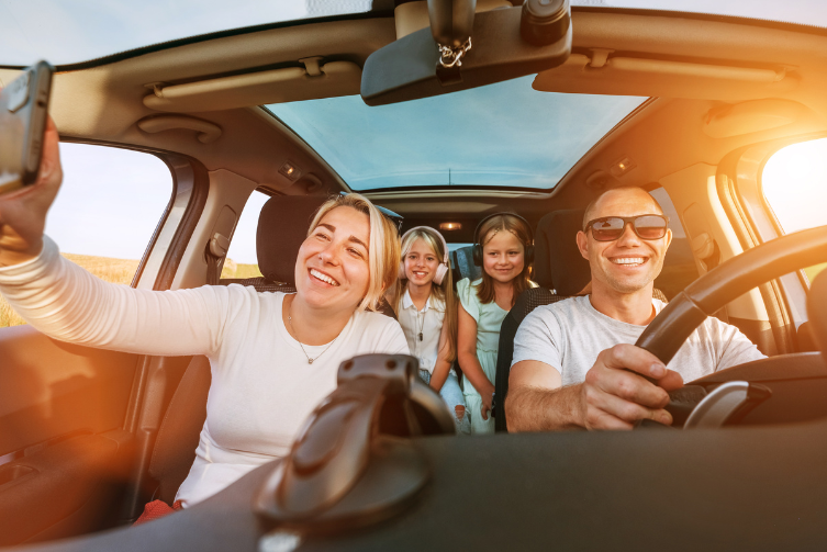 A father, mother and two young girls inside of a car. They pose for a photo from mom's phone camera.