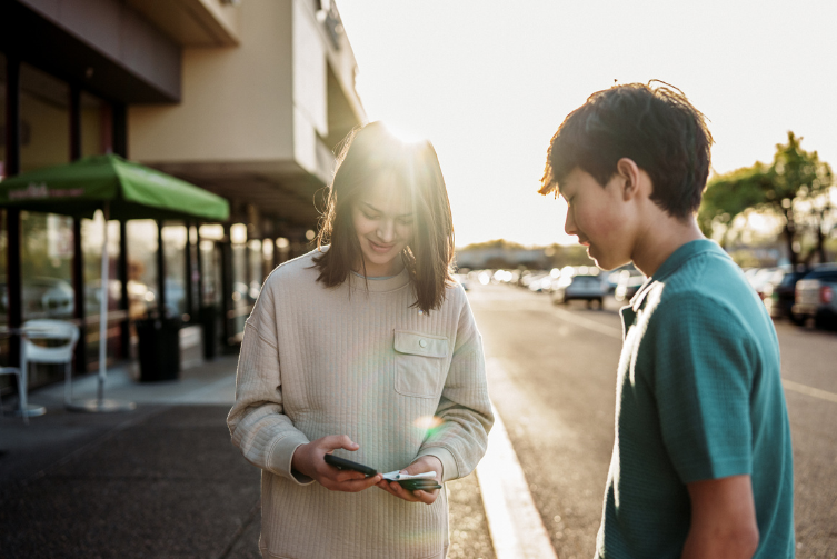 A teen girl uses her phone to manage finances with teen boy standing nearby