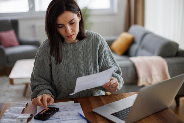 A woman checks her finances at her desk with a calculator and laptop