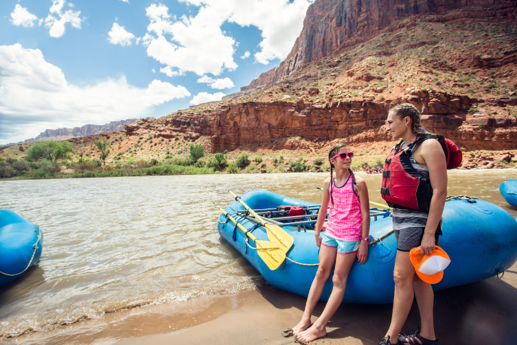 A woman and a girl lean against a raft alongside a desert river.