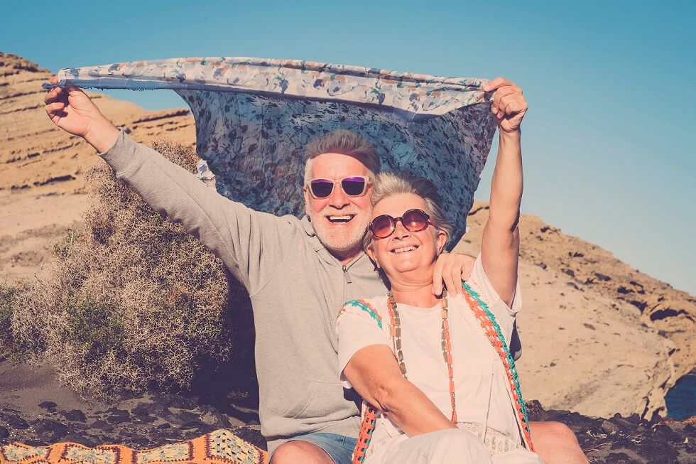 Older couple smiling in the sun, wearing sunglasses, holding up scarf.