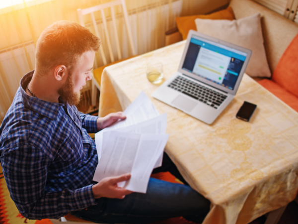 Man holds paperwork in his hands with his laptop nearby